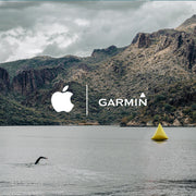 Scenic view of a swimmer in open water mode using a GPS-enabled swim tracking system, with a mountainous backdrop and a yellow buoy in the water. Logos of Garmin and Apple are displayed, indicating compatibility with these devices.